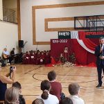 Mężczyzna w średnim wieku trzymający czerwoną różę w ręku ubrany w granatowy garnitur przemawia do mikrofonu na sali gimnastycznej.