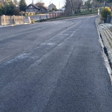 Rozległy asfaltowy plac. parking. W tle widać drogę, krzewy, domy oraz drzewa.