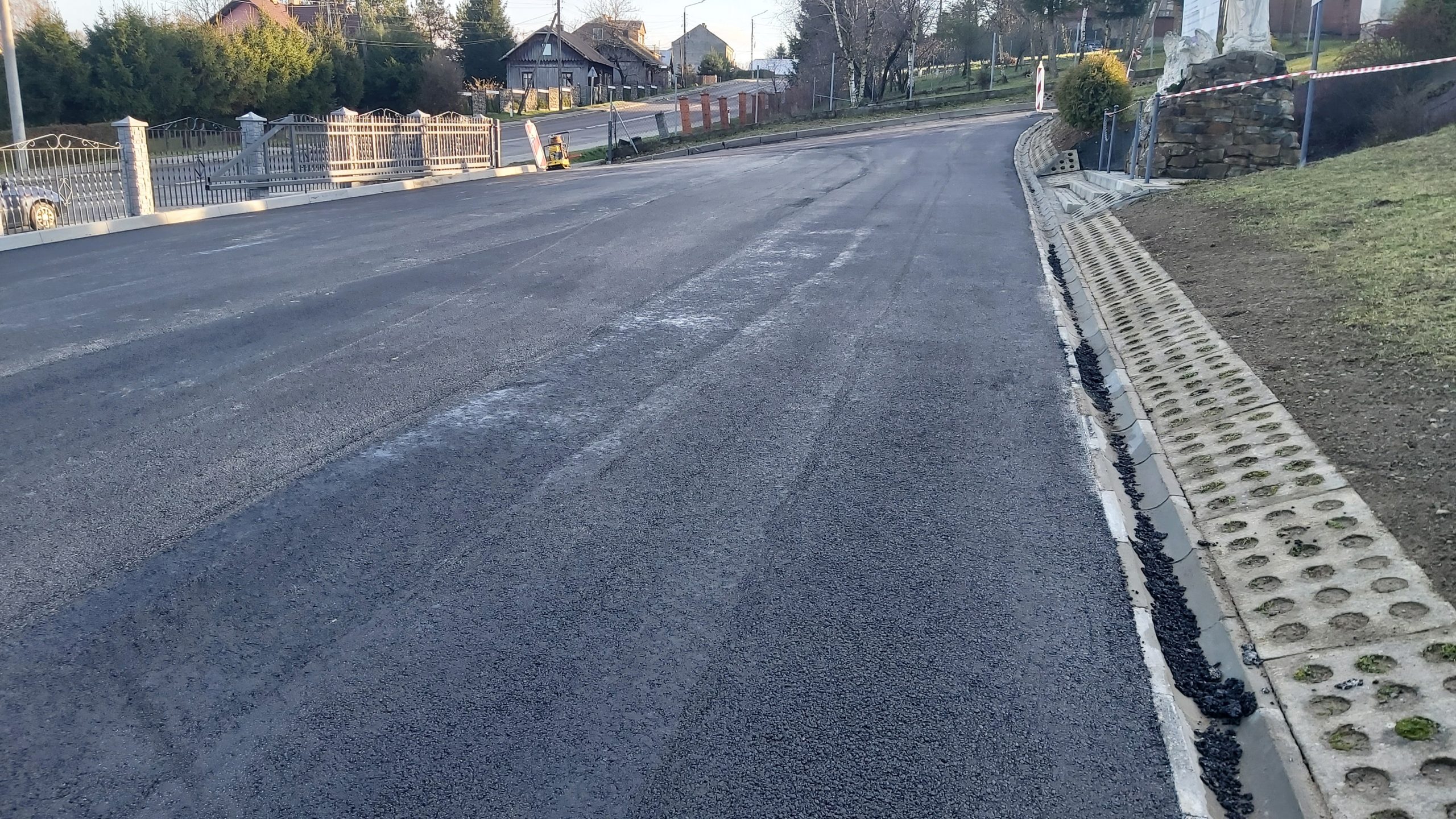 Rozległy asfaltowy plac. parking. W tle widać drogę, krzewy, domy oraz drzewa.