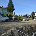 Remontowany plac na którym widać rozłożony kliniec, sprzęt i maszyny budowlane oraz dwa samochody. W tle można dostrzec dom oraz drzewa i krzewy. Panuje piękna słoneczna pogoda.