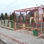 Przystanek drogowy przy chodniku z kostki brukowej. W tle domy i krzewy.