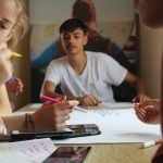 Młody chłopak w białej koszulce siedzi przy stole i rysuje na białych kartkach,.