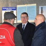 Trzech dorosłych mężczyzn rozmawiających przed ścianą z ogłoszeniami. Jeden z nich ubrany jest w garnitur, drugi w strój z napisem sędzia, a trzeci w ciemnogranatową kurtkę.