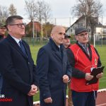 Pięciu dorosłych mężczyzn stojących na stadionie. W tle widzimy samochody, domy i drzewa.