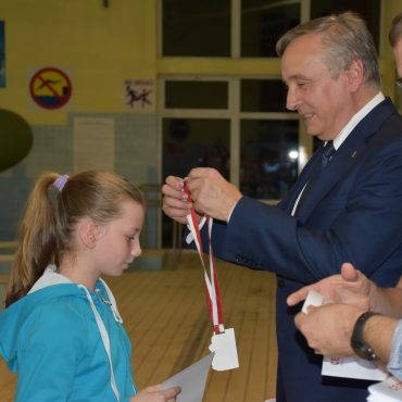 Dorosły mężczyzna w garniturze wręcza dziewczynce biało-czerwoną zawieszkę.