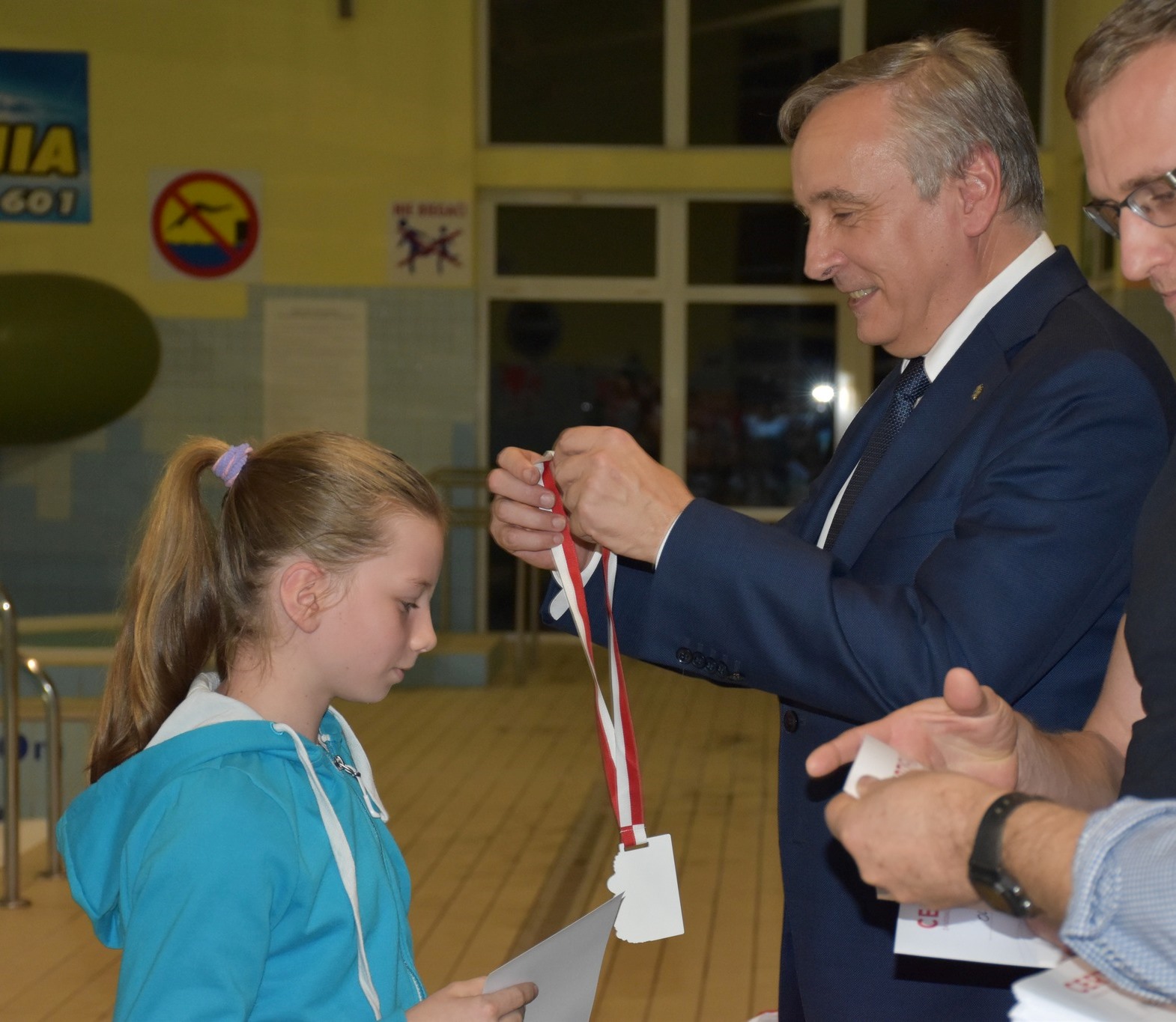 Dorosły mężczyzna w garniturze wręcza dziewczynce biało-czerwoną zawieszkę.