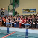 Duża grupa dzieci pozująca do zdjęcia przed basenem w rozległym pomieszczeniu. Po prawej strony grupy stoi dwóch dorosłych mężczyn.