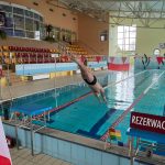 Basen pływacki w dużym pomieszczeniu, Widać mężczyznę w stroju pływackim wskakującego do wody.