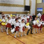 Grupa dzieci ubrana w białe bluzki i czerwone spódnice oraz chłopcy ubrani w stroje żołnierskie tańczą z pomponami żółtymi i białym na sali gimnastycznej.