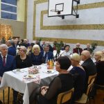 Grupa osób starszych mężczyzn i kobiet siedzi przy stoliku nakrytym białym obrusem.