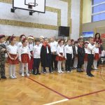 Grupa dzieci w strojach biało czerownych przedstawiaja na sali gimnastycznej.