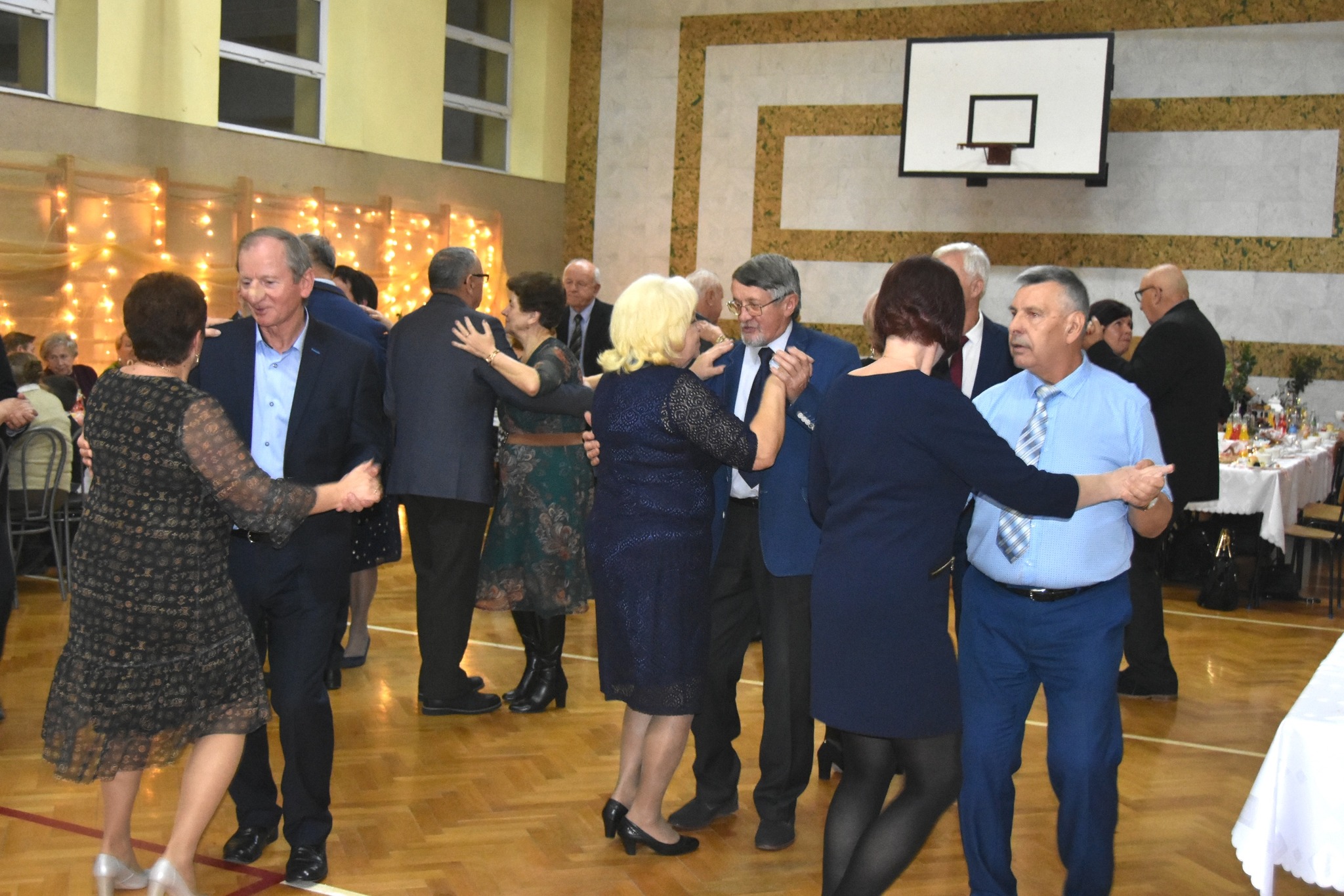 Grupa straszych osób tańczy na sali gimnastycznej.