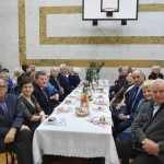 Grupa osób starszych mężczyzn i kobiet siedzi przy stoliku nakrytym białym obrusem.