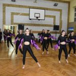 Grupa dziewczyn ubranych w czarne legnisy i koszulki z frędzlami fioletowymi tańczy na sali gimnastycznej.