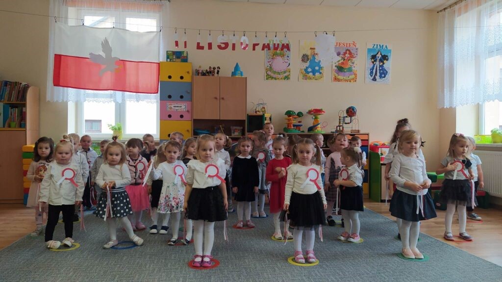 Grupa dzieci ubrana w odświętne stroje, z kotelionami biało czerownymi tańczy w sali szkolnej.