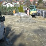 Remontowany plac na którym widać rozłożony kliniec, sprzęt i maszyny budowlane oraz trzech mężczyzn ubranych w strój roboczy. W tle można dostrzec ulicę, domy oraz drzewa i krzewy. Panuje piękna słoneczna pogoda.