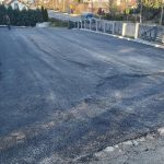 Rozległy asfaltowy plac. parking. W tle widać drogę, krzewy, domy oraz drzewa.