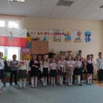 Grupa dzieci ubrana w odświętne stroje stoi na dywanie w sali szkolnej i recytuje wiersze. Za nimi na ścianie wisi duża flaga biało-czerwona