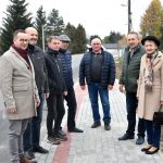 Szesciu dorosłych mężczyzn i kobieta stojący w cieplejszych, jesiennych strojach na chodniku z kostki brukowej.