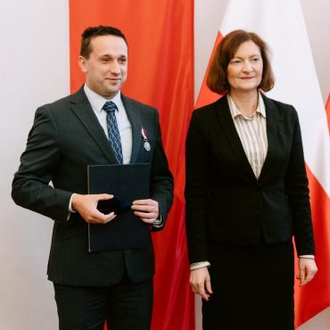 Młody mężczyzna w garniturze stoi z kobietą o krótkich włosach w czarnym garniturze, za nimi flagi biało-czerwone.