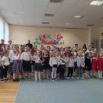 Grupa kilkudziesięciu dzieci ubranych w odświętne stroje spiewa hymn państwowy.