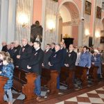 Grupa osób i dzieci stojąca przy kościelnych ławkach i patrząca uważnie przed siebie. Wszyscy są odświętnie ubrani.