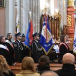 Kilka pocztów sztandarowych stojąca w kościele. W tle widać osoby siedzące na ławce.