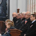 Grupa osób i dzieci siedząca na kościelnych ławkach i patrząca uważnie przed siebie. Wszyscy są odświętnie ubrani.