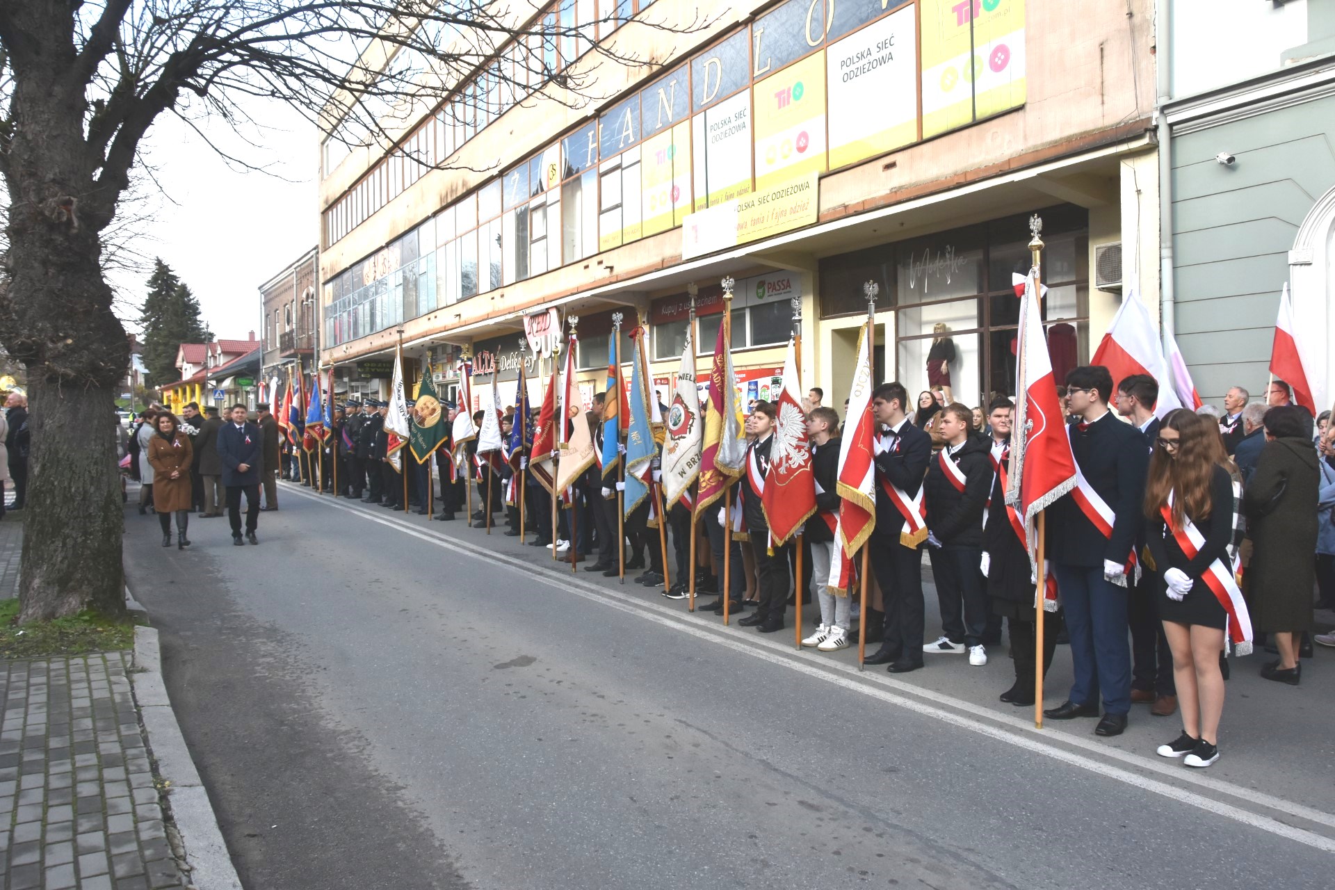 Rząd pocztów sztandarowych stojąca w długim rzędzie na ulicy. W tle widać gmach rozległego budynku.