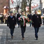 Trzech dorosłych mężczyzn w garniturze idących przed siebie. Idący w środku trzyma wieniec kwiatów z biało-czerwoną wstęgą. Z tyłu widać tłum ludzi.