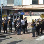 Orkiestra dęta w mundurach grająca na instrumentach. W tle droga i rozległy budynek.