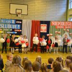 Grupa dzieci przed kurtyna z motywami patriotycznymi wymachuje biało-czerwonymi pomponami. Wszyscy znajdują się na sali gimnastycznej. Dzieci ogląda publiczność złożona głównie z uczniów.