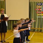 Grupa dzieci uroczysnie ślubująca z wyciągniętymi prawymi dłoniami. Wszyscy znajdują się na sali gimnastycznej. W tle widać dwie dorosłe kobiety oraz starsza dziewczynkę.