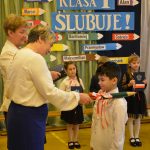 Kobieta dokonująca uroczystego aktu pasowania na młodym uczniu ubranym w odświętny, biały strój. Obok kobiety stoi druga, przekazująca uczniom dyplomy.