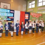 Grupa dzieci przed kurtyną z motywami patriotycznymi trzyma w rękach ozdobne dyplomy.