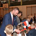 Dwóch dorosłych mężczyn na sali gimnastycznej wręcza uczniom upominki i biało czerwone flagi.