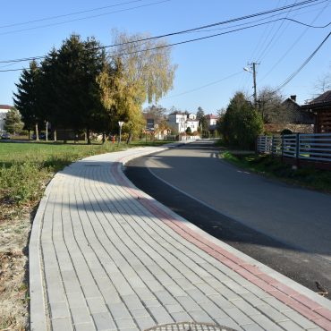 Chodnik z kostek brukowych oraz droga asfaltowa. W tle lampy, domy, budynki gospodarcze i krzewy.