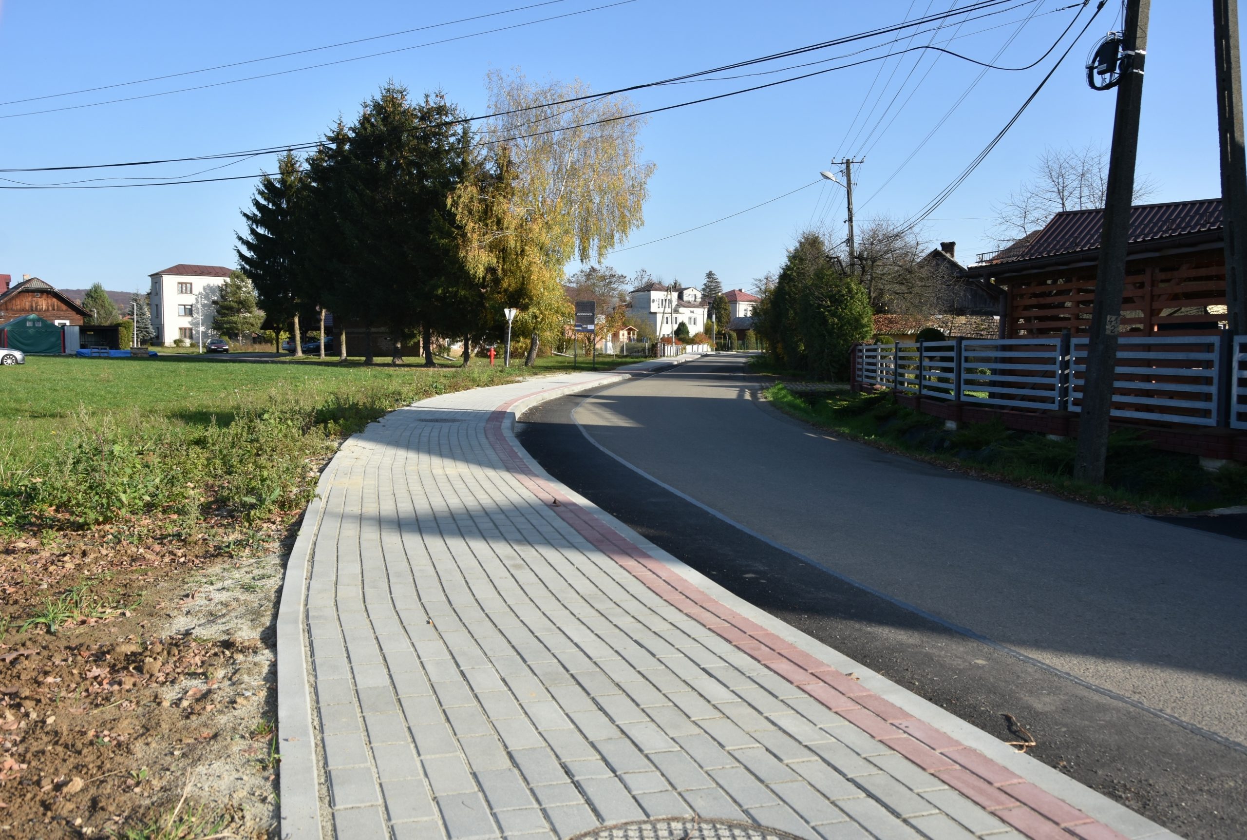 Chodnik z kostek brukowych oraz droga asfaltowa. W tle lampy, domy, budynki gospodarcze i krzewy.