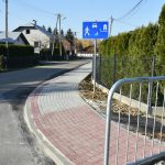 Chodnik z kostek brukowych oraz droga asfaltowa. Przy chodniku znajduje się niebiesko-biały znak. W tle domy, budynki gospodarcze i krzewy.