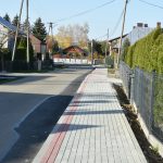 Chodnik z kostek brukowych oraz droga asfaltowa. W tle domy, budynki gospodarcze i krzewy.
