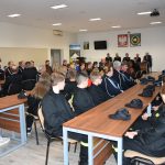 Grupa osób różnej płci i wieku zasiadajaca przy podłużnych stolach na rozległej sali. Osoby są ubrane głównie w stroje strażackie.