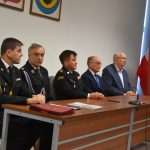 Grupa pięciu mężczyzn zasiadająca przy podłużnym stole na rozległej sali. Osoby te ubrane są w mundury galowe oraz garnitury.