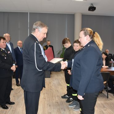 Mężczyzna w średnim wieku w mundurze gratulujący kobiecie w mundurze strażackim. Znajdują się w rozległym pomieszczeniu. Za nimi znajduje się grupa osób odświętnie ubranych w rożnym wieku.