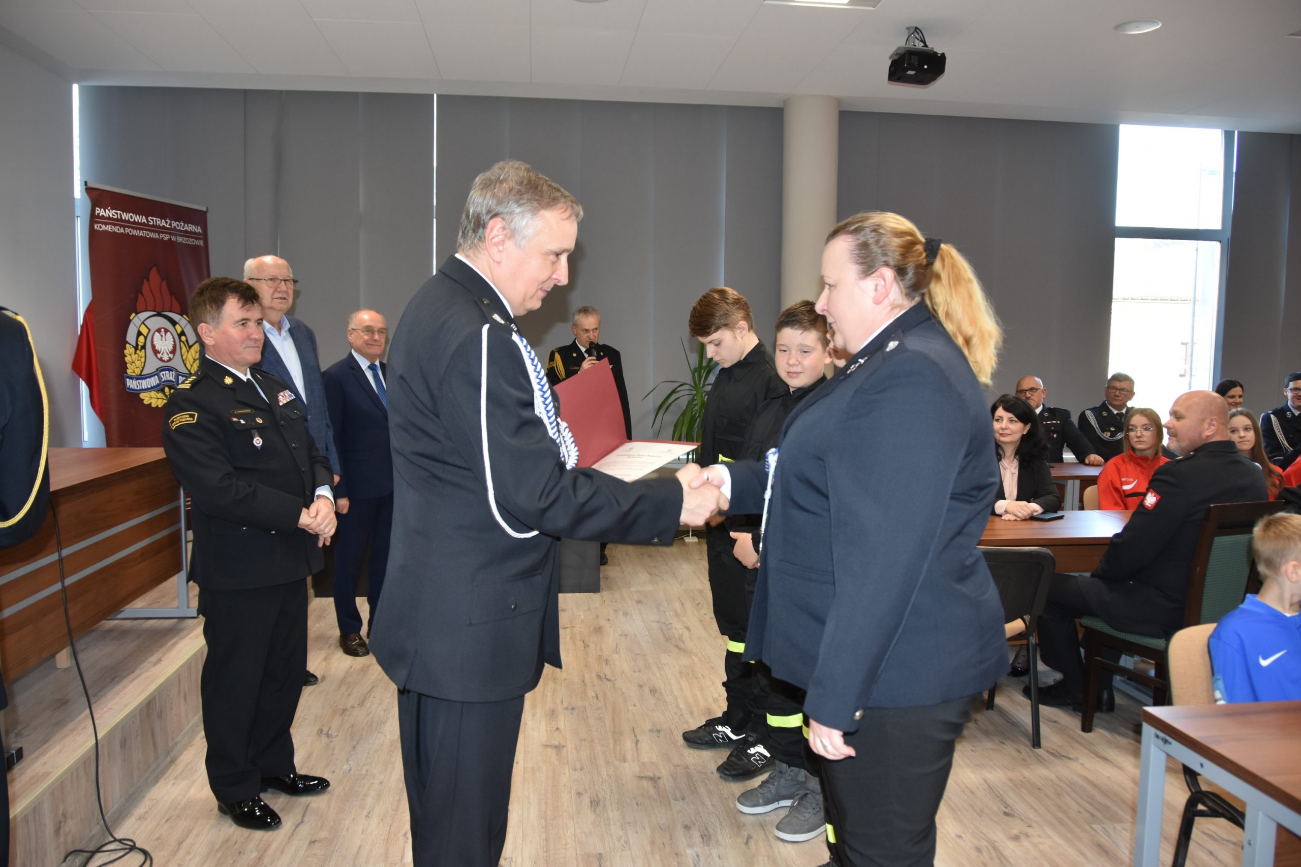 Mężczyzna w średnim wieku w mundurze gratulujący kobiecie w mundurze strażackim. Znajdują się w rozległym pomieszczeniu. Za nimi znajduje się grupa osób odświętnie ubranych w rożnym wieku.