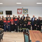 Duża grupa osób obojga płci pozująca do zdjęcia na wielkiej sali konferencyjnej. Na ścianie widać herby i godła oraz telewizor i obrazy. W te znajdują się dwa rolapy.