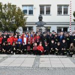 Duża grupa osób obojga płci pozująca do zdjęcia przed gmachem urzędu. Oosby są ubrane w stoje strażackie oraz oficjalne ubiory urzędowe. W tle widać duże krzewy oraz pomnik - popiersie Józefa Piłsudskiego.