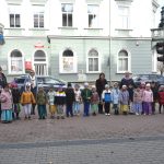 Grupa dzieci i kilku dorosłych kobiet w jesiennych strojach, stojacy w grupie do zdjęcia. w Tle gmach dużego, zabytkowego budynku oraz przejście dla pieszych i kościół.