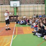 Grupa dzieci siedzi na sali gimnastycznej, przed nimi stoi młody mężczyzna który do nich przemawia.