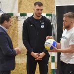 Stoi trzech młodych mężczyzn na sali gimnastycznej. Jeden w sportowej koszulce podaje drugiemu w garniturze białą piłkę do siatkówkii. Za nimi stoi bramka.