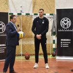 Trzech mężczyzn stoi na sali gimnastycznej. Jedn z garniturze trzyma piłke do siatkówki oraz mikrofon do którego przemawia.